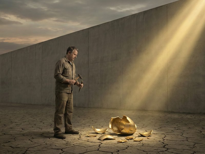 Muž stojící před metaforickou zdí dokonalosti s rozbitou zlatou nádobou u nohou.